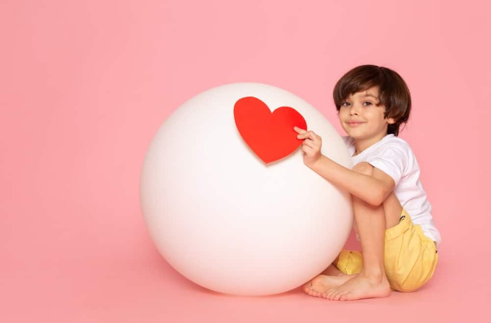 Un ragazzo sorride mentre posa un cuore rosso su una grande sfera bianca su sfondo rosa