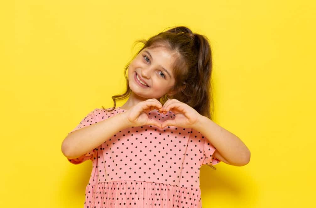 Una ragazza allegra con un abito a pois forma un cuore con le mani su uno sfondo giallo