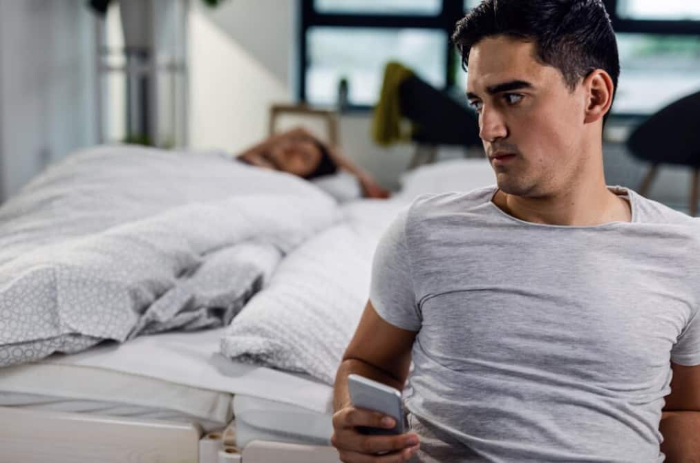 Un uomo seduto sul letto che guarda il telefono con una donna che dorme sullo sfondo