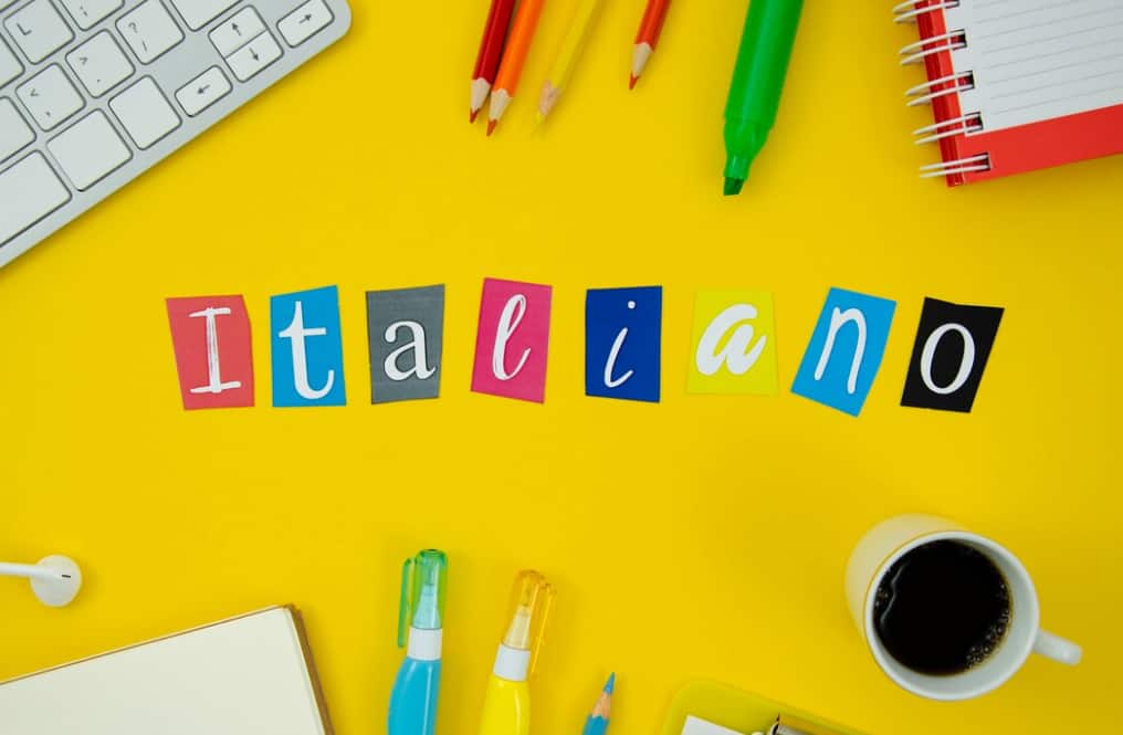 Lettere colorate che compongono l'ortografia 'Italiano' su sfondo giallo con articoli di cancelleria